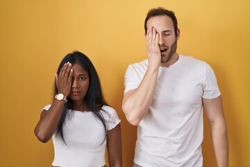 Interracial couple standing over yellow background yawning tired covering half face, eye and mouth with hand. face hurts in pain.