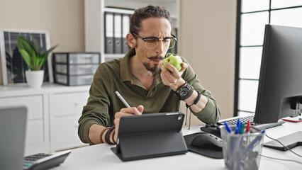 Young hispanic man business worker writing on touchpad eating apple at office