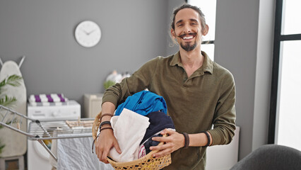 Young hispanic man smiling confident holding basket with clothes at laundry room