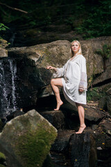 A smiling happy pregnant girl in a light vintage dress, with blond short hair, stands against the backdrop of picturesque rocks near a waterfall in the forest.