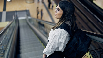 Young beautiful hispanic woman standing on escalator wearing backpack in subway station of Madrid