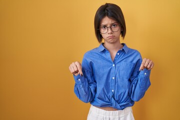 Young girl standing over yellow background pointing down looking sad and upset, indicating...