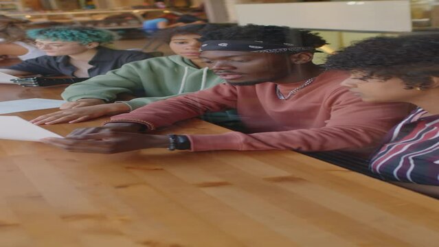 Vertical shot of of gen Z diverse friends sitting in cafe behind glass window, reading menu and discussing dishes before ordering food in cafe
