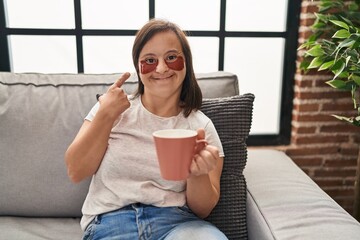 Hispanic girl with down syndrome sitting on the sofa drinking a coffee at home smiling happy pointing with hand and finger