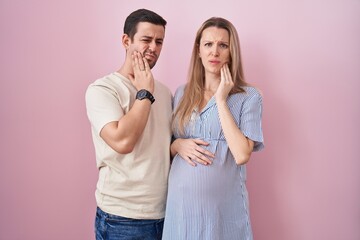 Young couple expecting a baby standing over pink background touching mouth with hand with painful...