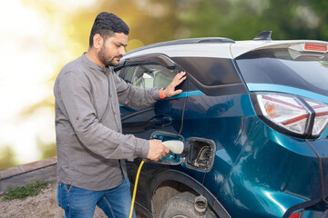 Indian man attaching power cable into socket to charge his modern electric car at outdoor charging station. Electric vehicle Recharging battery charging port. Renewable energy or green energy.