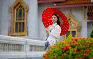 A young woman dressed in retro Thai dress stands holding a red umbrella inside Wat Benchamabophit Dusitwanaram Rajaworawihan, looking at the flower field and smiling at the camera.