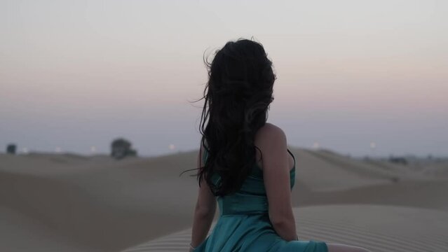 Young woman with black hair sitting barefoot in a long blue evening dress on the sand dunes of the desert