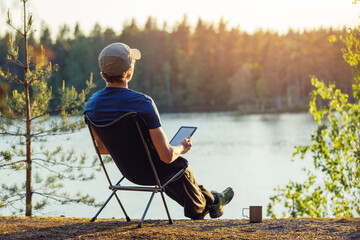 A man is reading an e-book on the shore of a forest lake at sunset in the evening.
