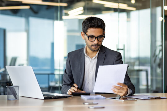 Serious concentrated hispanic businessman financier on paperwork inside office, man in business suit at workplace reviewing and reading papers, contracts and accounts reports