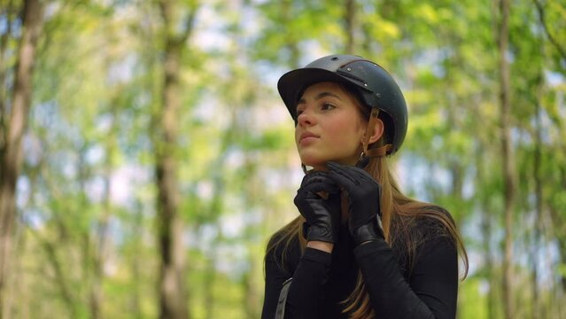Smiling young female equestrian fastening helmet looking away. Portrait of confident Caucasian beautiful woman putting on uniform in sunny park forest outdoors