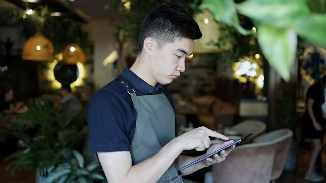 Serious cafe employee wearing dark green pinafore taking online order via portable tablet. Attractive restaurant manager in uniform conveying online order via internet to delivery service.