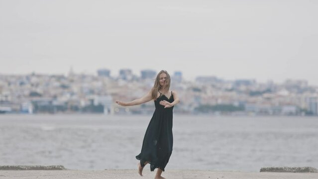 An adult cute woman in black dress dancing on the pier on the background view of european town