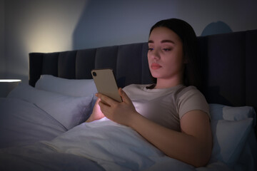 Young woman using modern smartphone in bed at night. Internet addiction