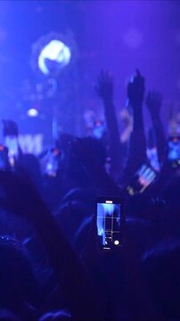 Vertical video slow motion of unrecognizable fans dancing at a concert or festival party. Silhouettes of concert crowd in front of bright flashing strobe stage lights. High quality footage