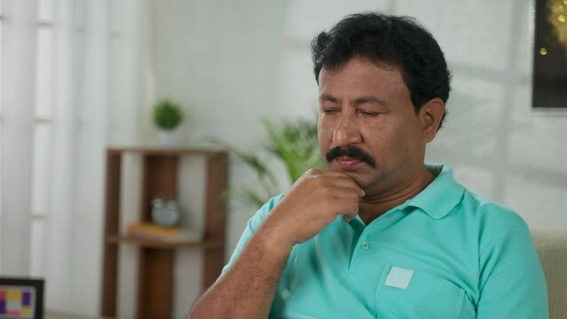 Thoughtful worried indian middled aged man deep thinking while sitting on sofa at home - concept of loneliness, financial problems and mental illness