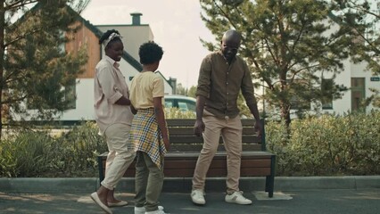 Stab shot of cheerful Black family of three sitting down on wooden bench outside while walking together on warm sunny day
