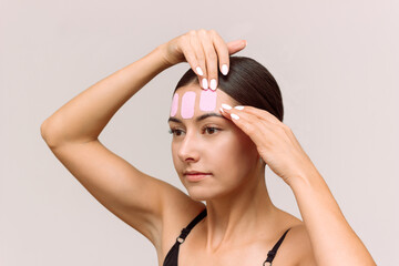 A young Caucasian woman puts pink kinesio tape on her face to reduce wrinkles