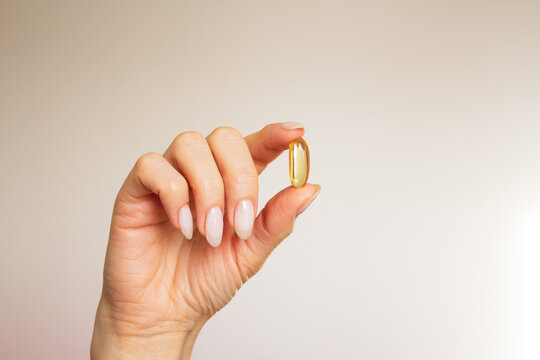  woman's hand holdind with her fingers a soft yellow gel capsule close-up on a beige background with sunlight.