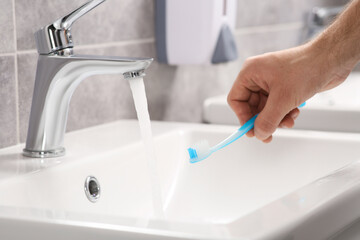 Man holding toothbrush near flowing water above sink in bathroom, closeup