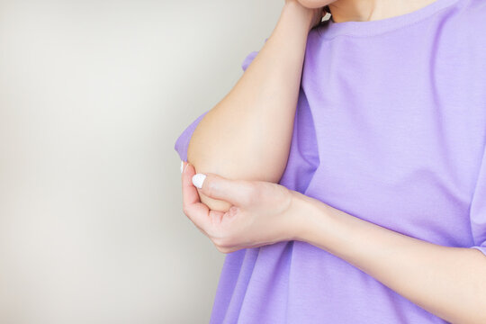 lose-up photo of a young woman in a purple T-shirt holding the elbow joint, experiencing pain from arthritis, elbow injury, isolated on a light background
