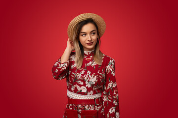 Stylish young lady touching straw hat and looking away in red studio