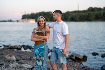 Fototapeta premium Cheerful young teenage lovers near the riverbank. She is feeling confident smiling holding skateboard and he is looking at her.