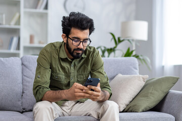 Serious thinking man sitting on sofa in living room at home, Indian man reading news on phone, using app on smartphone, browsing internet pages.