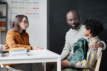 Young teacher talking to father of boy about his behavior while they sitting in classroom after...