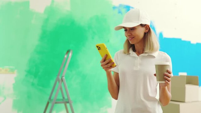 Female worker standing, texting with clients during painting the wall and drinking coffee. Woman in special uniform scrolling apps in her phone on the break. People and technology concept