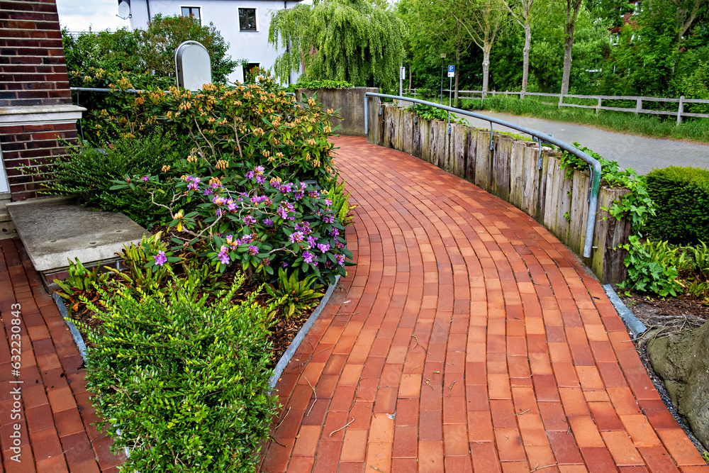 Poster red brick path with flowers and bushes on either side of it and white building in the background in 