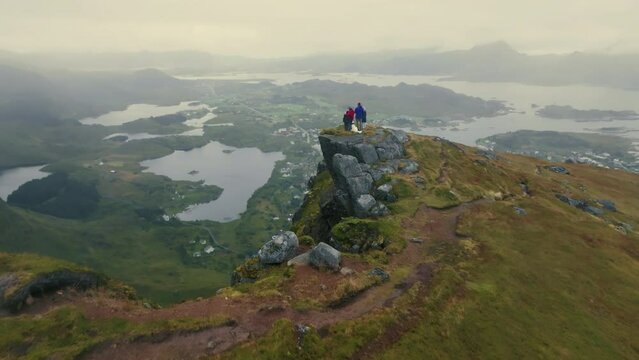Drone flies over a young family with a white dog standing on the edge of a fjord towards an epic panorama of a town in the Lofoten Islands