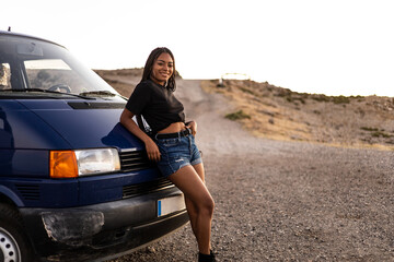 A Latina girl with braided hair in casual clothes is leaning on a camper van while looking at the...