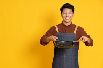 Young Asian man chef wearing kitchen apron cooking and holding pan and spatula isolated on yellow background