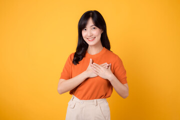 Happy young Asian woman in her 30s, wearing an orange shirt, holds hands on chest on yellow...