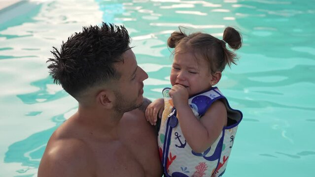 Video of a little girl with children's life jacket crying in the arms of her young father while they take a bath inside the water of the pool at home.