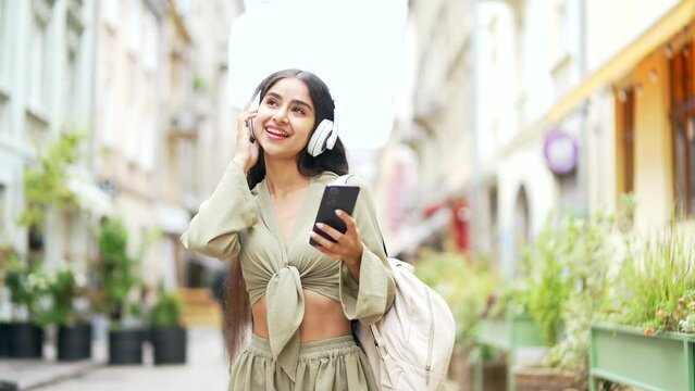 Joyful Young Indian Arabian Woman Listening To Music In Wireless Headphones Using A Smartphone Phone While Walk On A City Street Outdoor Cheerful Female Girl Dancing Concept Of Happiness And Lifestyle