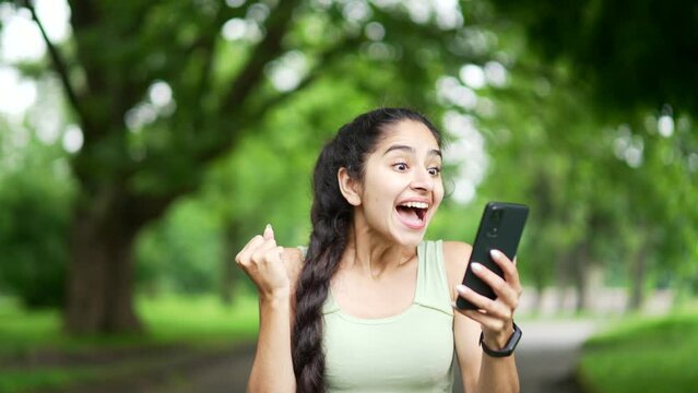 Happy excited young sports woman checking results of training while looking at smartphone in urban city park. Smiling female happy with good successful workout results or great news on mobile phone