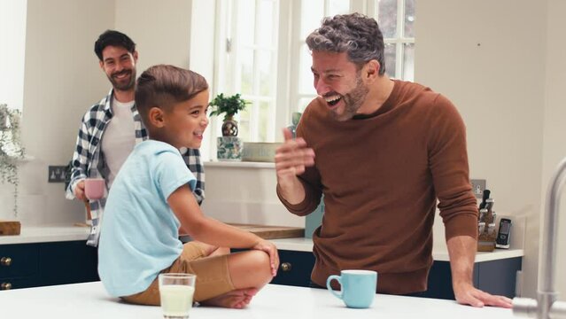 Same sex family with two Dads in kitchen at home with son sitting on counter giving parent high five - shot in slow motion