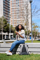 Young happy African American woman sitting in city park using mobile phone shopping online or texting messages. Smiling female student holding cellphone chatting in sunny campus area, vertical.