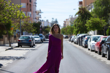 Fototapeta premium Young beautiful blonde woman dressed in purple dress walks along a large lonely avenue among the stopped cars. The woman makes different body expressions while parading like a model.