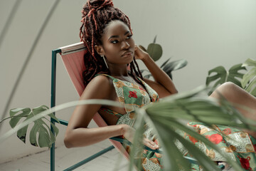 Charming African woman, wearing a multicolor ornament dress, sitting in a hammock chair surrounded...