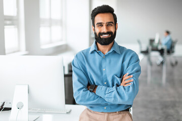 Successful young indian businessman in shirt standing with folded arms near desk, smiling at...