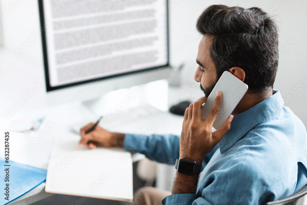 Poster indian businessman having conversation on smartphone, sitting in front of computer monitor, taking n