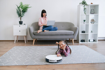 Focus on well-rounded robot vacuum cleaner being placed on floor of living room by cute child in...