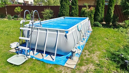 The gray rectangular frame pool stands on a grassy lawn. The surface of the water is covered with a floating awning. Nearby there are thuas and flowers in flowerbeds. The pool is made of PVC