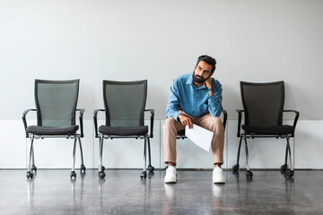 Bored indian man with CV tired of waiting for employment interview at office lobby, sitting on...