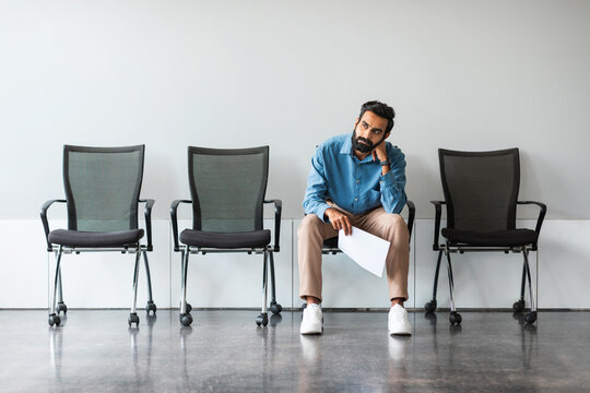Bored indian man with CV tired of waiting for employment interview at office lobby, sitting on chair, free space