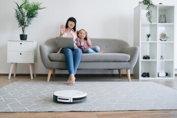 Obraz premium Selective focus of modern robotic vacuum keeping floor tidy while female owners starting video call on gadget. Mother-daughter duo enjoying online communication while wireless appliance doing job.