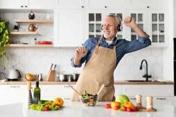 Positive Senior Man In Headphones Listening Music While Cooking Food In Kitchen